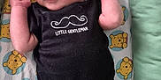 Thomas is registered to the contest to win money with this photo: baby, infant, onesie, black_clothing, mustache_graphic, text_on_clothing, teddy_bear_pattern, blanket, lying_down, curious_expression, arms_up, face, head, person, soft_fabric, bed, pajamas, cute, indoor, child