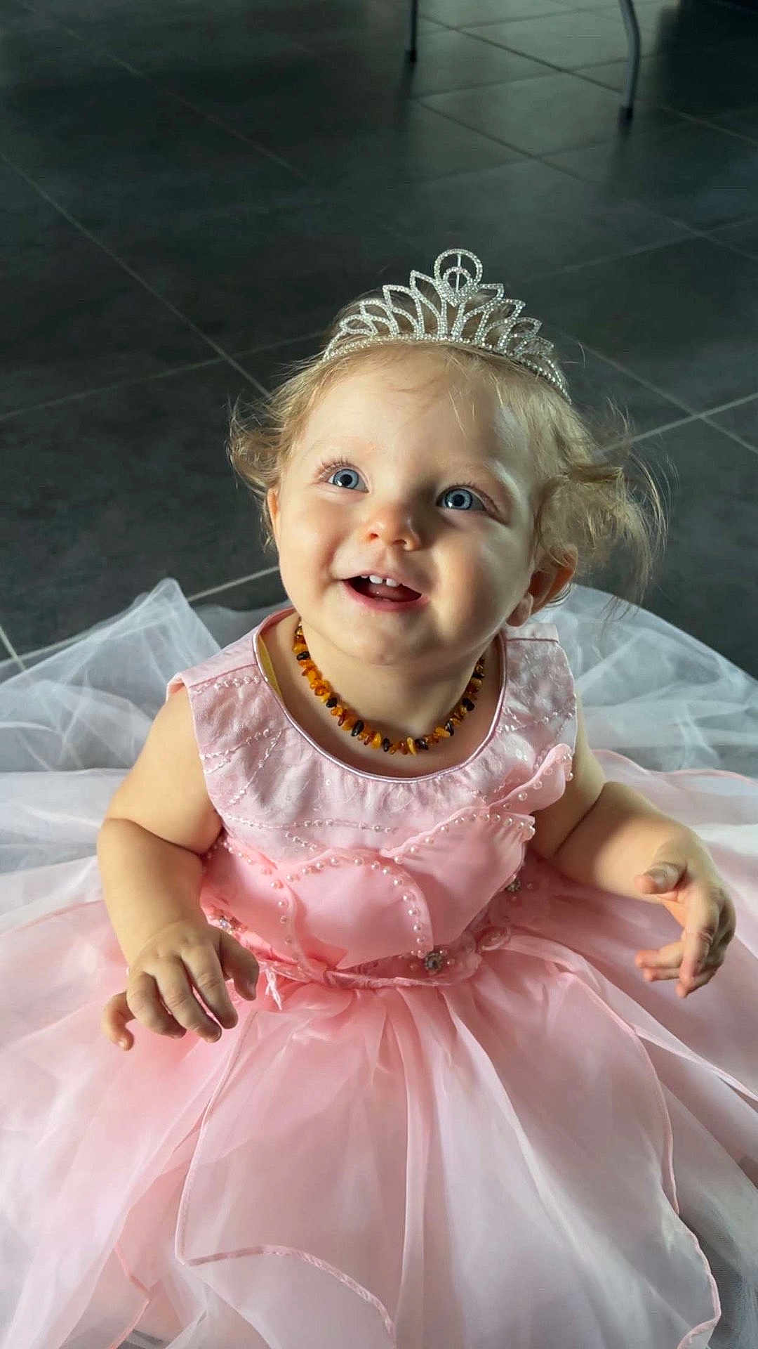 Louhann participe au concours pour gagner de l'argent avec cette photo : child, toddler, princess, tiara, pink_dress, smiling, happy, blue_eyes, curly_hair, necklace, floor, tiles, indoor, portrait, cute, baby, sitting, dress, celebration, joyful