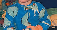 John is registered to the contest to win money with this photo: bed, bedroom, blue_pajamas, child, clothing, cute, dinosaur_pattern, expression, face, furniture, holding, home, indoor, person, portrait, sitting, sleepwear, toddler, toy, young_child