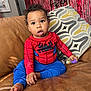 barefoot, blue_pants, casual_pose, child, cozy, curious_expression, curly_hair, decorative_pillow, home_decor, indoor, leather_couch, living_room, portrait, purple_toy, red_shirt, red_streamers, sitting, spiderman_outfit, toddler, young_child