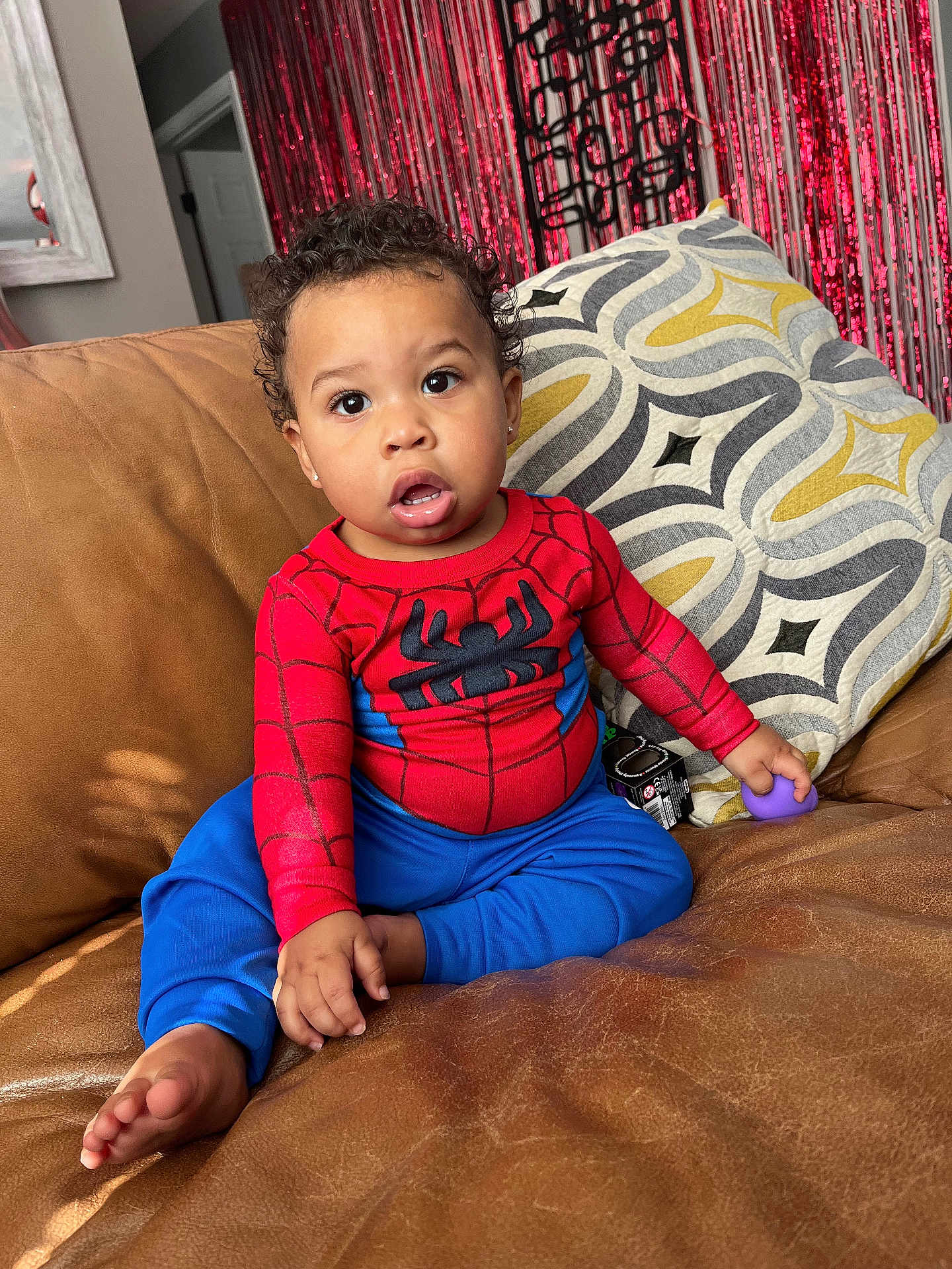 Karmelo joined the competition — help win amazing prizes! barefoot, blue_pants, casual_pose, child, cozy, curious_expression, curly_hair, decorative_pillow, home_decor, indoor, leather_couch, living_room, portrait, purple_toy, red_shirt, red_streamers, sitting, spiderman_outfit, toddler, young_child