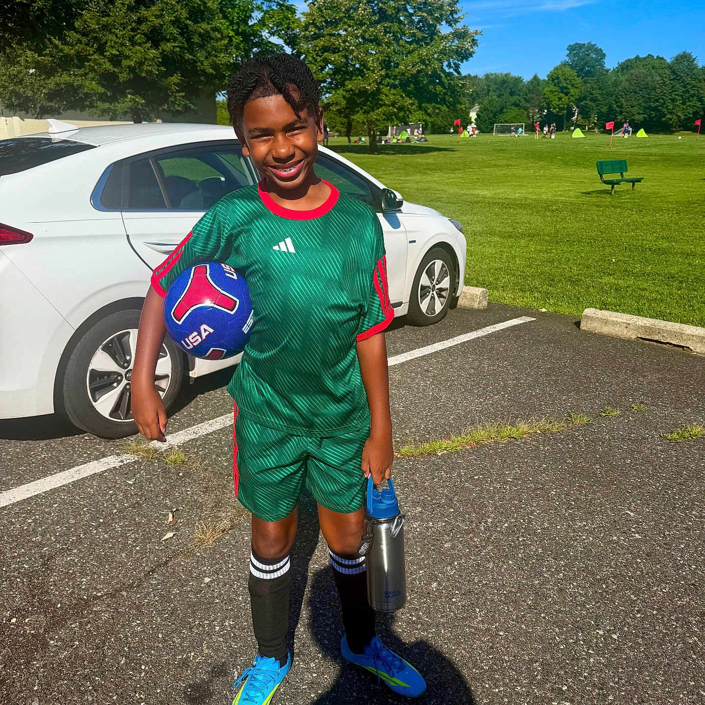 Josh is registered to the contest to win money with this photo: child, soccer_ball, sportswear, water_bottle, car, parking_lot, green_field, trees, blue_sky, sunny_day, soccer_cleats, grass, person, outdoor, smiling, shorts, socks, athlete, recreation, activity