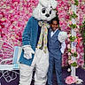 Josh joined the competition — help win amazing prizes! child, boy, mascot, bunny, costume, flower_wall, pink_flowers, suit, bow_tie, glasses, smiling, posing, celebration, event, bench, purple_floor, decorations, white_fur, happy, portrait