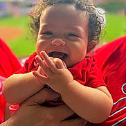 Lisa is registered to the contest to win money with this photo: adult_hand, baby, bow, child, closeup, curly_hair, cute, green_background, hand, happy, holding, infant, joyful, laughing, outdoor, person, portrait, red_clothing, smiling, stadium