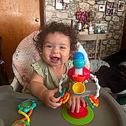 Lisa is registered to the contest to win money with this photo: baby, child, curly_hair, high_chair, toy, smiling, happy, indoor, family_photos, wall_decor, furniture, table, colorful, plastic, hand, clothing, person, portrait, cute, playtime