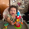 baby, child, curly_hair, high_chair, toy, smiling, happy, indoor, family_photos, wall_decor, furniture, table, colorful, plastic, hand, clothing, person, portrait, cute, playtime