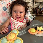 Lisa joined the competition — help win amazing prizes! baby, child, curly_hair, smiling, high_chair, bib, toy, sun_toy, bee_toy, indoor, face, happy, infant, clothing, blanket, table, person, cute, portrait, playful