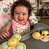 baby, child, curly_hair, smiling, high_chair, bib, toy, sun_toy, bee_toy, indoor, face, happy, infant, clothing, blanket, table, person, cute, portrait, playful