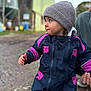 toddler, child, outdoor, snowsuit, hat, gray_hat, navy_clothing, pink_clothing, person, hand, gravel, farm_building, blurred_background, cold_weather, winter_clothing, portrait, side_view, curious_expression, supporting_hand, daylight