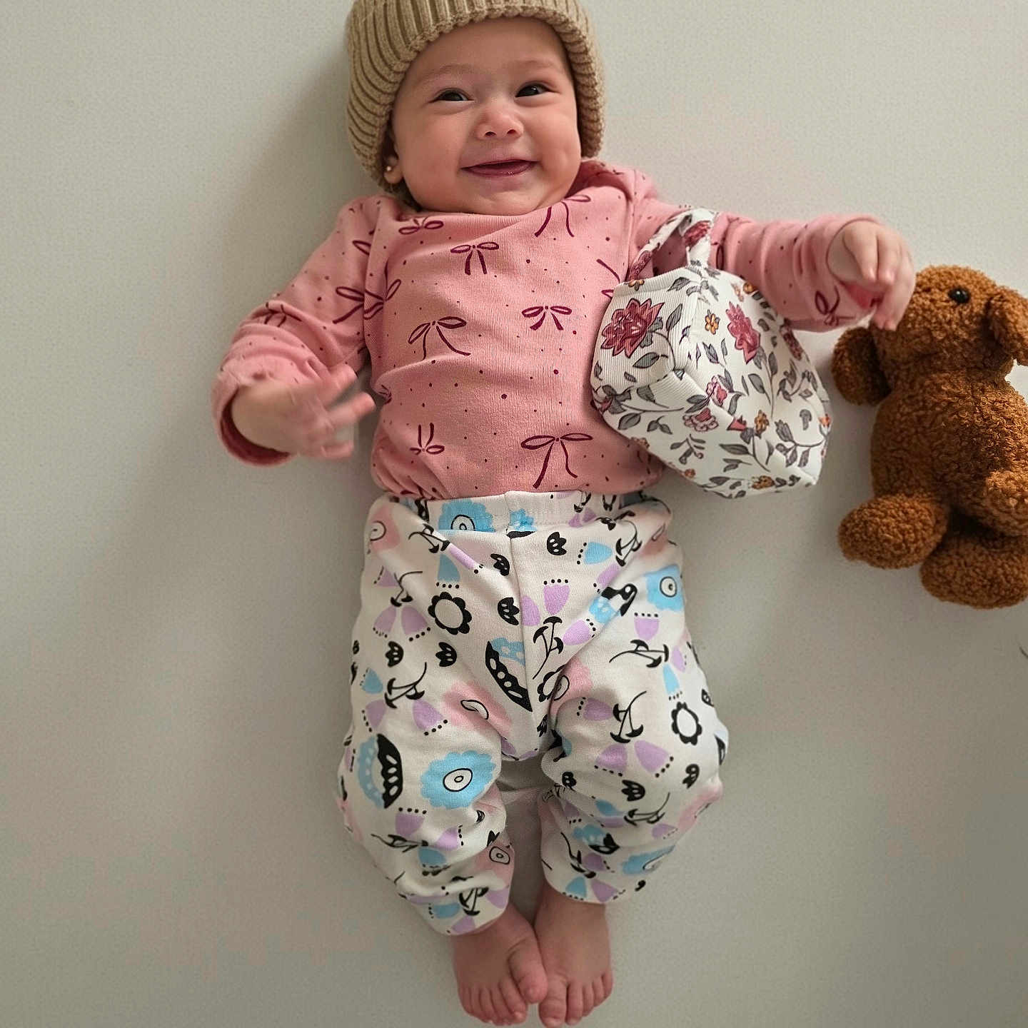 Allison joined the competition — help win amazing prizes! baby, child, smiling, portrait, indoors, lying_down, knit_hat, pink_clothing, pajamas, patterned_clothing, teddy_bear, stuffed_animal, toy, floral_hat, infant, face, hands, feet, wall, ai_generated_content