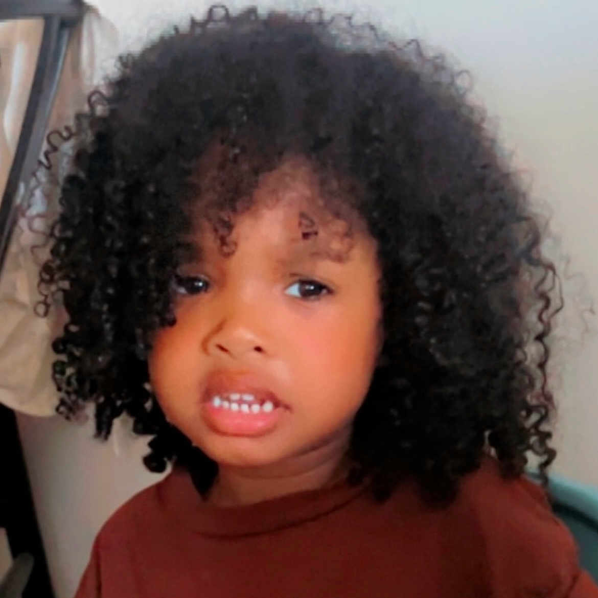 Isaac participe au concours pour gagner de l'argent avec cette photo : brown_shirt, chair, child, close_up, clothing, curly_hair, cute, dark_hair, expression, eyes, face, headshot, indoor, mouth, nose, person, portrait, teeth, wall, young_child