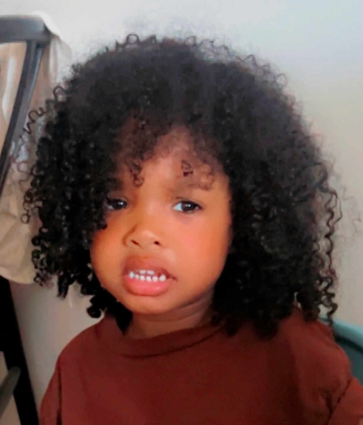 Isaac participe au concours pour gagner de l'argent avec cette photo : child, young_child, portrait, curly_hair, dark_hair, face, teeth, expression, clothing, brown_shirt, indoor, close_up, headshot, eyes, mouth, nose, chair, wall, person, cute