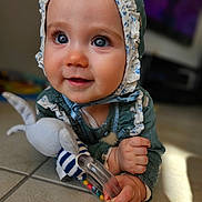 Enola a rejoint le concours — aidez-le/la à gagner de superbes lots ! baby, child, blue_eyes, bonnet, toy, rattle, floor, tile, hand, face, infant, cute, smiling, indoors, person, closeup, clothing, playing, expression, portrait