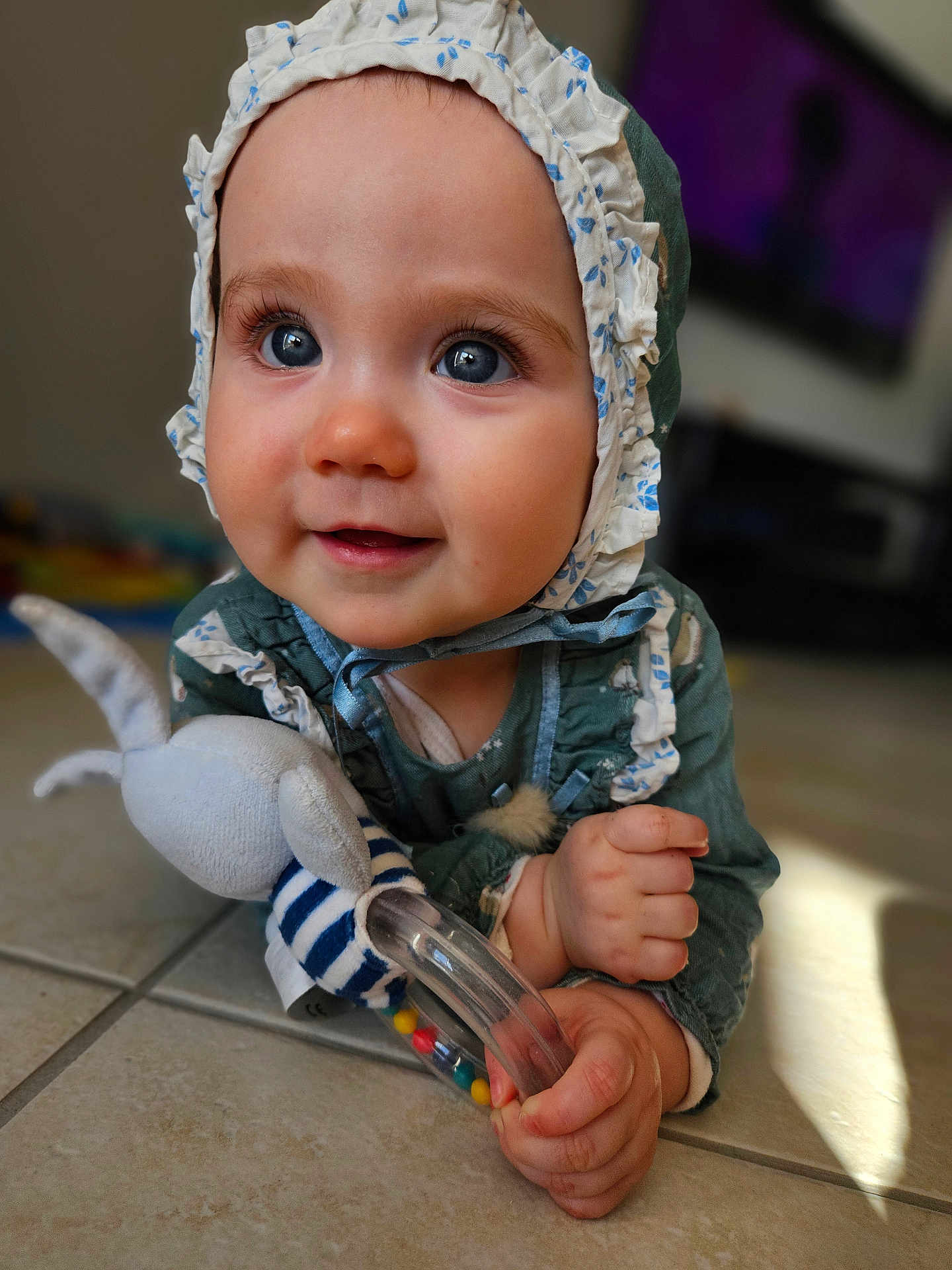 Enola a rejoint le concours — aidez-le/la à gagner de superbes lots ! baby, child, blue_eyes, bonnet, toy, rattle, floor, tile, hand, face, infant, cute, smiling, indoors, person, closeup, clothing, playing, expression, portrait