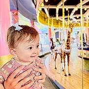Liya joined the competition — help win amazing prizes! baby, child, hands, carousel, giraffe, amusement_ride, indoor, lights, smile, hair_bow, pink_dress, patterned_clothing, person, portrait, happy, playful, colorful, fun, family, holding