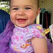 Maliya is registered to the contest to win money with this photo: apparel, baby, cartoon_characters, cheerful, child, closeup, cute, daylight, face, happy, infant, natural_light, outdoor, person, porch, portrait, purple_dress, sitting, smiling, tulle