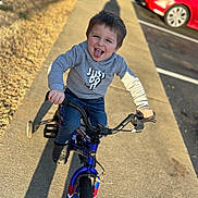 Landyn is registered to the contest to win money with this photo: child, boy, bicycle, training_wheels, sidewalk, gray_hoodie, jeans, playful, tongue_out, sunlight, shadow, parking_lot, cars, outdoor, happy, smiling, person, concrete, casual_clothing, daytime