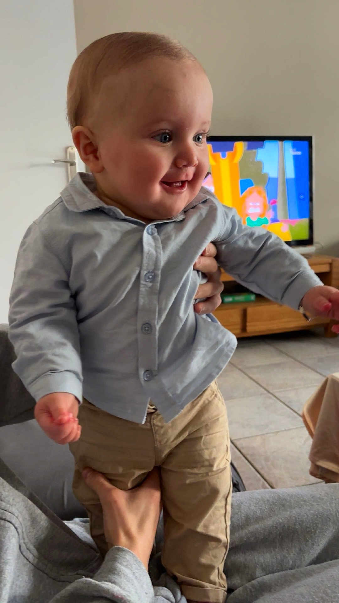 Charly a rejoint le concours — aidez-le/la à gagner de superbes lots ! baby, child, toddler, smile, button_up_shirt, pants, standing, adult_hand, living_room, television, cartoon, wooden_furniture, tile_floor, couch, portrait, cute, happy, indoor, face, person