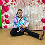 boots, brown_dog, casual_clothing, cross_legged, cute, decorations, dog, floor, floor_tile, glasses, heart_backdrop, indoor, indoor_lighting, matching_outfits, person, pet, playful_expression, posing, sitting, tie_dye_hoodie