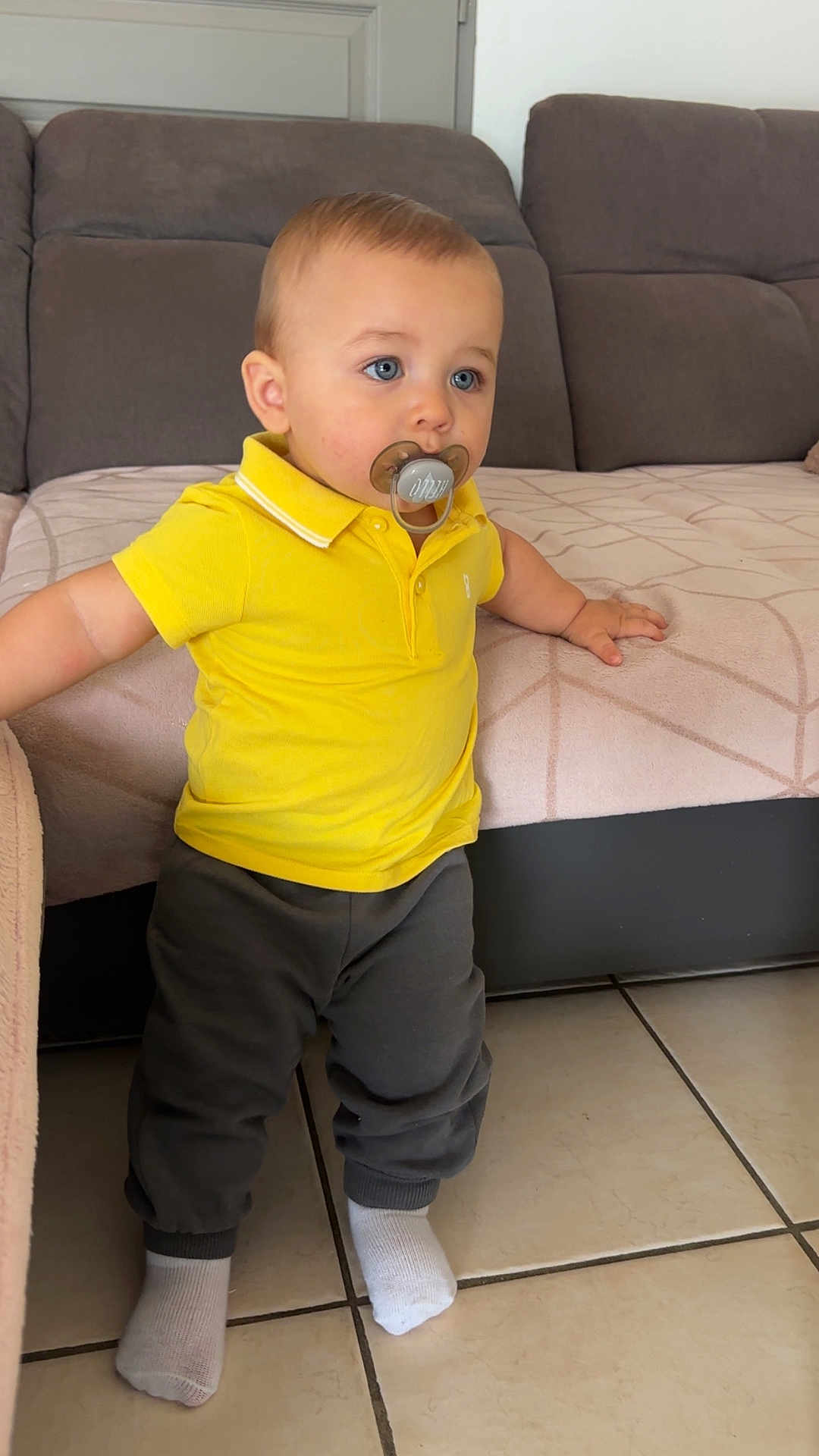 Khyronn participe au concours pour gagner de l'argent avec cette photo : toddler, child, pacifier, yellow_polo, pants, socks, blue_eyes, standing, indoor, couch, chair, tile_floor, baby, person, young_child, home, furniture, casual_clothing, curious, portrait