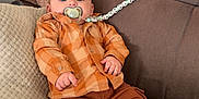 Khyronn a rejoint le concours — aidez-le/la à gagner de superbes lots ! baby, infant, pacifier, orange_shirt, brown_pants, white_socks, couch, pillows, beaded_pacifier_clip, indoor, child, cute, relaxed, plaid, clothing, furniture, person, portrait, sitting, home