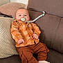 baby, infant, pacifier, orange_shirt, brown_pants, white_socks, couch, pillows, beaded_pacifier_clip, indoor, child, cute, relaxed, plaid, clothing, furniture, person, portrait, sitting, home