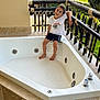 child, boy, jacuzzi, balcony, ocean, palm_trees, vacation, smiling, outdoor, summer, relaxation, white_tub, shorts, tshirt, railing, tiles, sunlight, nature, happy, leisure