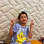 child, boy, pikachu, tshirt, peace_sign, smile, shorts, sitting, indoor, patterned_wall, orange_surface, happy, casual_clothing, portrait, curly_hair, playful, young, person, comfortable, relaxed