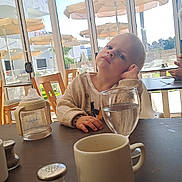 Noé participe au concours pour gagner de l'argent avec cette photo : child, toddler, indoor, table, glass, water, baby_bottle, cup, salt_shaker, window, umbrella, palm_tree, chair, daylight, casual_clothing, person, expression, resting_head, restaurant, bright