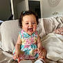 baby, child, smiling, sitting, dress, curly_hair, indoor, couch, cushion, footwear, happy, person, face, home, furniture, colorful, patterned_clothing, cute, joyful, infant
