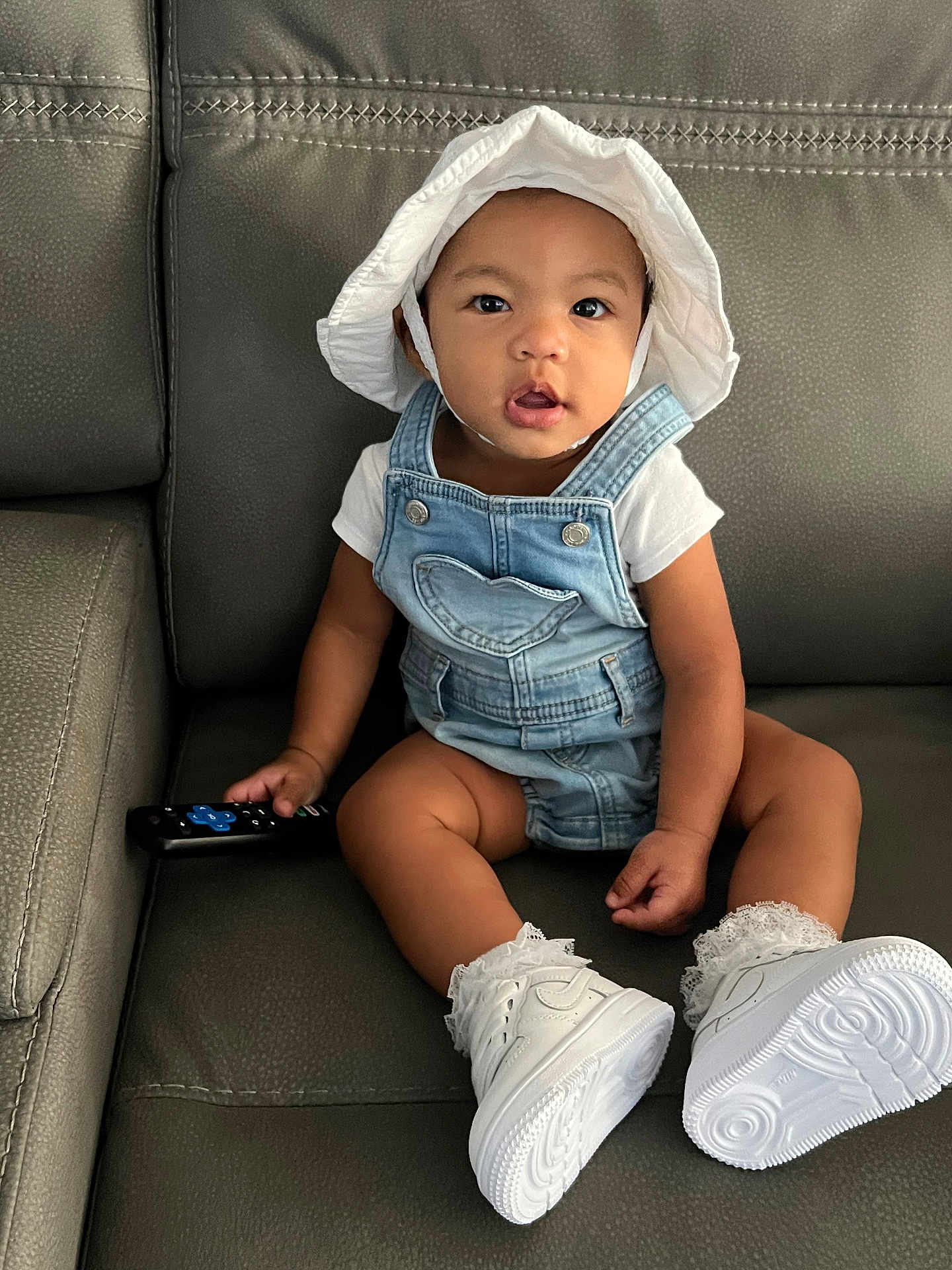 Maylee joined the competition — help win amazing prizes! baby, casual, child, clothing, couch, curious, denim_overalls, expression, face, footwear, hat, home, indoor, lace_socks, leather, person, remote_control, sitting, toddler, white_shoes