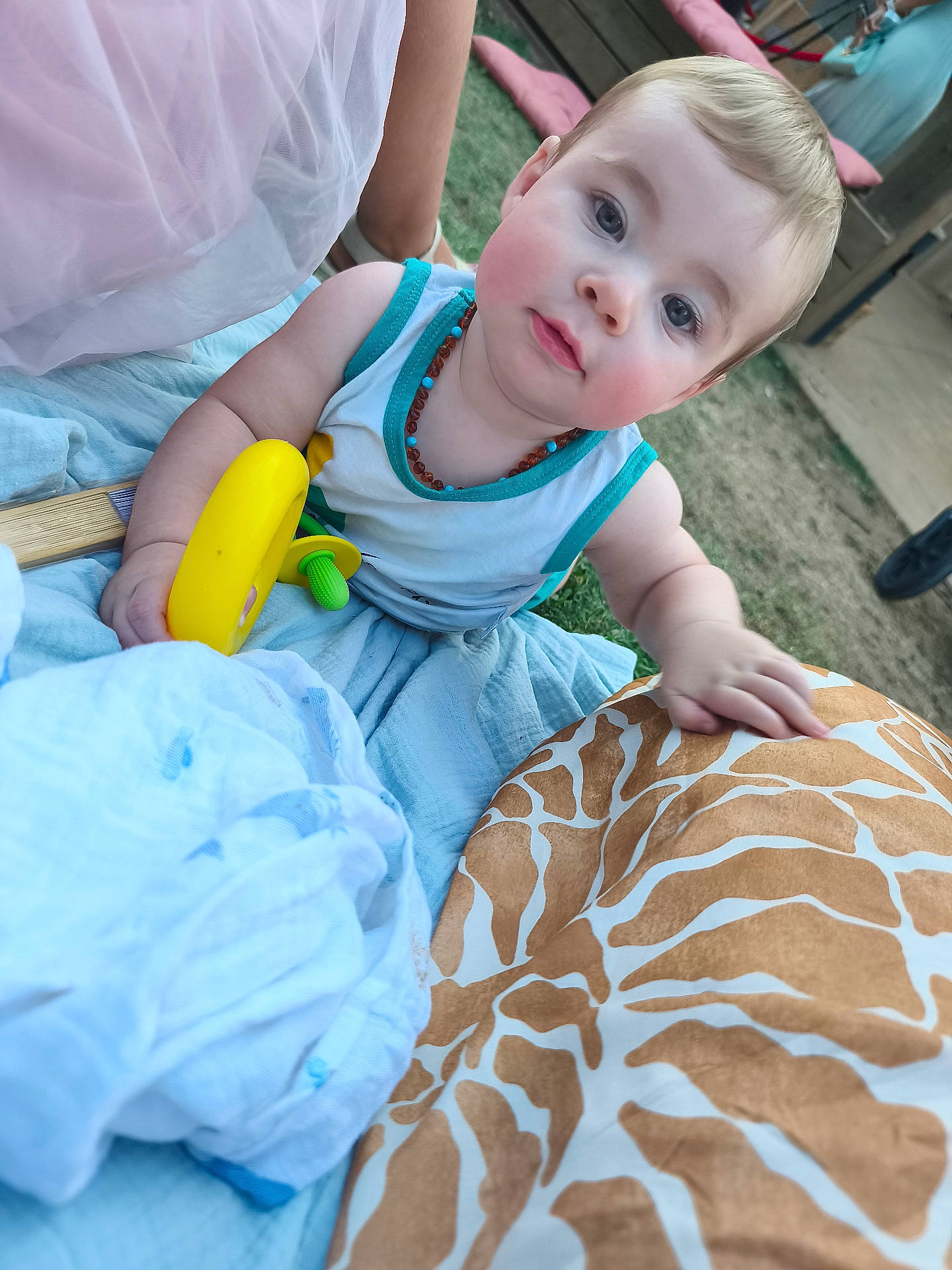 baby, blanket, blue_eyes, casual_clothes, child, curious, cute, daylight, family, gathering, grass, infant, necklace, outdoor, person, playtime, rosy_cheeks, soft_light, summer, toy