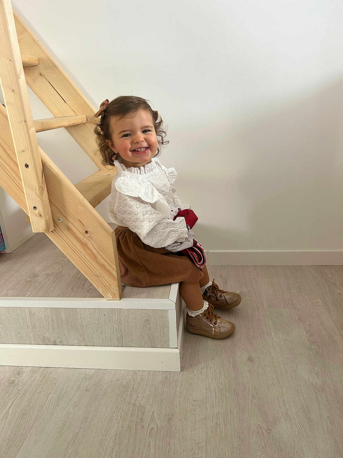 Ava participe au concours pour gagner de l'argent avec cette photo : child, toddler, smile, curly_hair, bow, white_blouse, brown_skirt, shoes, stairs, wood, floor, indoor, happy, sitting, person, cute, young_child, portrait, home, casual