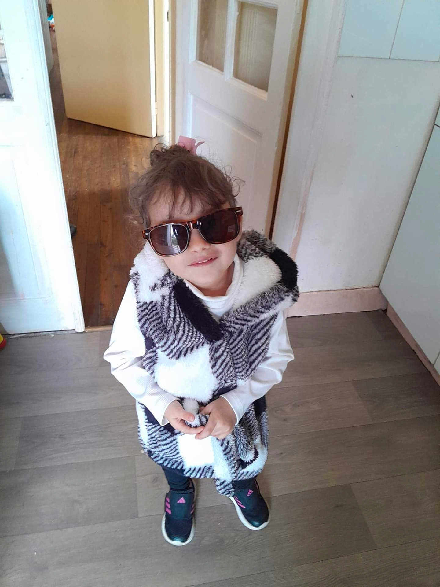 Mallaury participe au concours pour gagner de l'argent avec cette photo : black_pants, casual_clothing, child, cool, curly_hair, cute, door, fashion, footwear, home, indoor, person, pink_hair_tie, smile, sneakers, standing, sunglasses, vest, white_shirt, wooden_floor