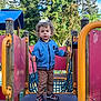 Ezio participe au concours pour gagner de l'argent avec cette photo : child, colorful, curly_hair, daylight, fun, greenery, jacket, outdoor, pants, person, play_area, play_structure, playground, rope_net, shoes, sky, standing, steering_wheel, toddler, trees