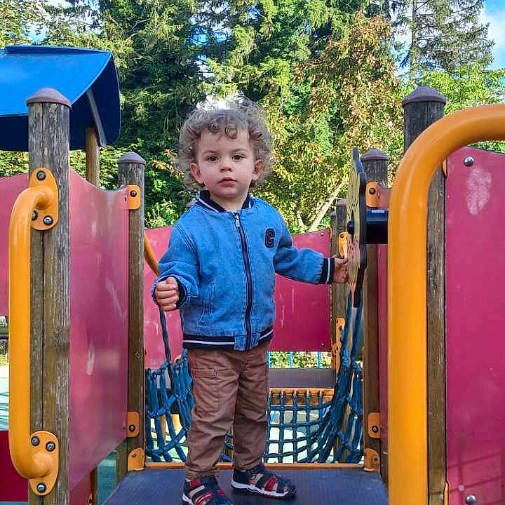 Ezio participe au concours pour gagner de l'argent avec cette photo : child, colorful, curly_hair, daylight, fun, greenery, jacket, outdoor, pants, person, play_area, play_structure, playground, rope_net, shoes, sky, standing, steering_wheel, toddler, trees