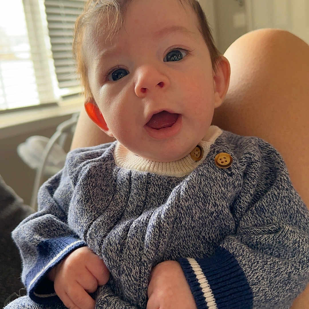Greyson is registered to the contest to win money with this photo: baby, blanket, bodypart, clothing, coat, couch, face, finger, furniture, hand, head, homedecor, jacket, knitwear, pants, person, photography, portrait, sitting, sweater