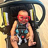 baby, heart_shaped_glasses, car_seat, smiling, child, infant, seatbelt, black_car_seat, happy, cute, indoor, wooden_floor, sitting, colorful_clothing, toddler, playful, adorable, young_child, glasses, seat