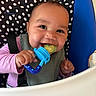 Milly is registered to the contest to win money with this photo: baby, child, high_chair, teething_toy, polka_dot, blue, green_food, happy, smiling, face, hand, feeding, infant, cute, indoors, clothing, pink_sleeves, seatbelt, closeup, portrait