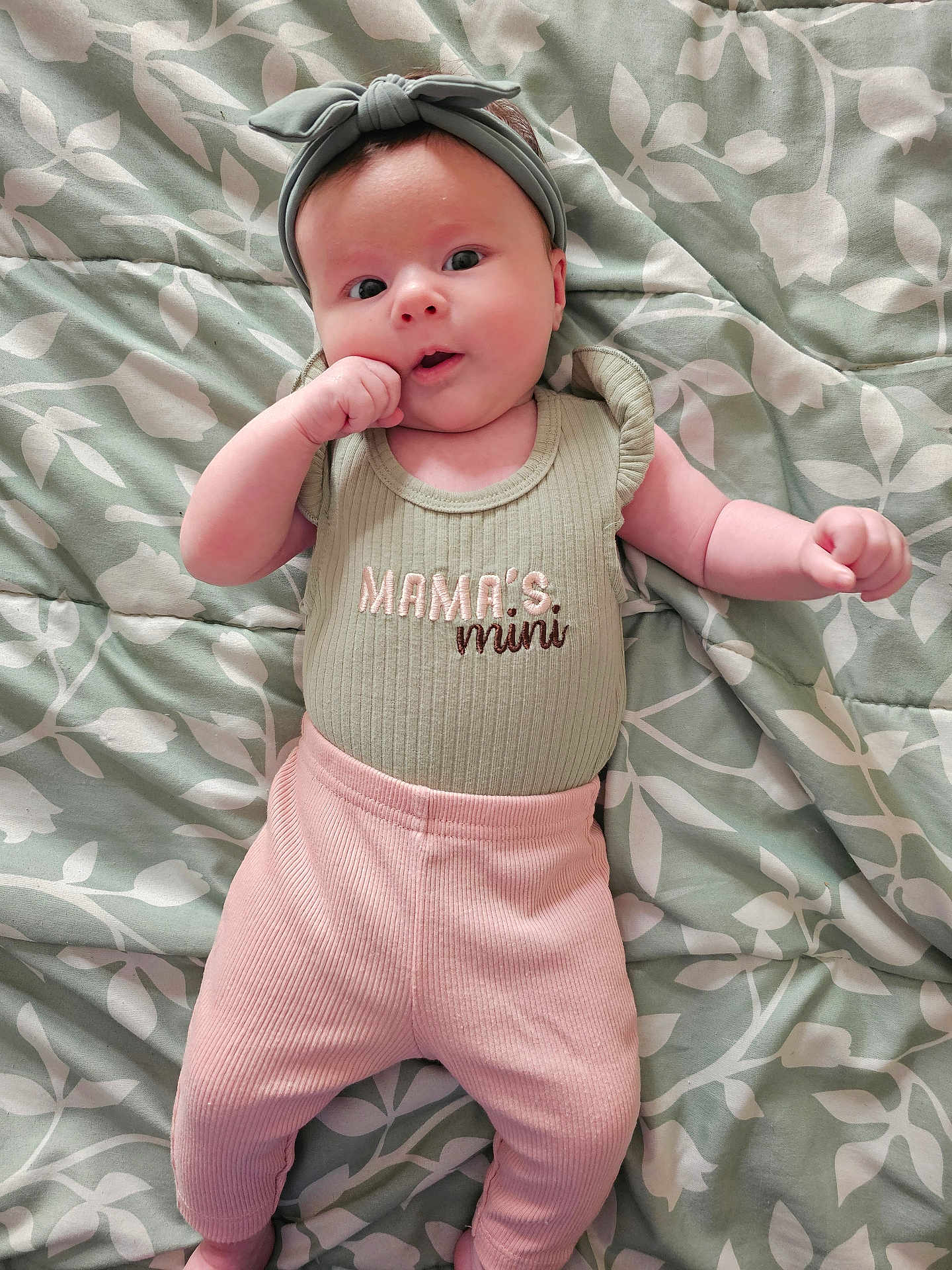 Lilith is registered to the contest to win money with this photo: baby, infant, headband, green, pink, clothing, blanket, pattern, leaf_pattern, person, cute, lying_down, hand_near_mouth, portrait, soft_lighting, indoors, child, apparel, fabric, expression