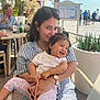 woman, toddler, child, smiling, outdoor, cafe, table, chair, sunglasses, striped_clothing, floral_pants, watch, plants, people, sunlight, happy, holding, portrait, casual, daytime