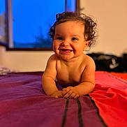 Lilou participe au concours pour gagner de l'argent avec cette photo : baby, child, smile, curly_hair, bed, indoor, window, evening, blue_sky, skin, portrait, happy, infant, lying_down, warm_lighting, room, colorful_bedspread, hands, face, cute