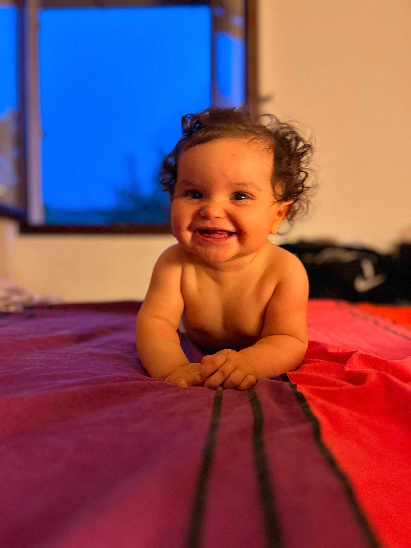 Lilou participe au concours pour gagner de l'argent avec cette photo : baby, child, smile, curly_hair, bed, indoor, window, evening, blue_sky, skin, portrait, happy, infant, lying_down, warm_lighting, room, colorful_bedspread, hands, face, cute
