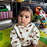 activity_center, baby, bangs_hair, brown_eyes, child, corduroy_pants, curious_expression, floral_pattern, gaming_controller, indoor, living_room, play_mat, portrait, shelf, sitting, soft_lighting, sweater, toddler, toy, tv