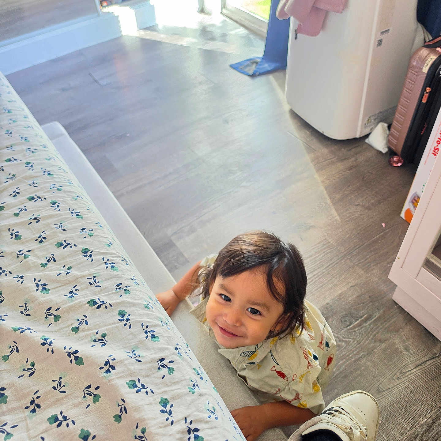 Franco is registered to the contest to win money with this photo: child, toddler, smiling, indoors, bedroom, bed, bedding, floral_pattern, floor, wood_floor, laundry, washing_machine, suitcase, window, natural_light, portrait, looking_at_camera, dark_hair, casual_clothing, family_home
