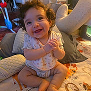 Jessie is registered to the contest to win money with this photo: baby, child, toddler, smiling, happy, curly_hair, portrait, sitting, indoors, playroom, play_mat, infant, baby_clothing, bare_feet, toys, cushion, furniture, warm_lighting, family_home, cute