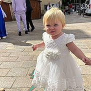 Grâce participe au concours pour gagner de l'argent avec cette photo : toddler, child, white_dress, blonde_hair, outdoor, sunny, cobblestone, street, people, building, adult, footwear, daylight, urban, sidewalk, smile, person, fashion, walking, casual