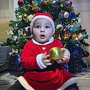 Esra a rejoint le concours — aidez-le/la à gagner de superbes lots ! baby, child, christmas_tree, ornament, holiday, festive, red_clothing, santa_hat, indoor, decorations, presents, sparkle, glitter, cute, celebration, seasonal, smiling, sitting, person, toy