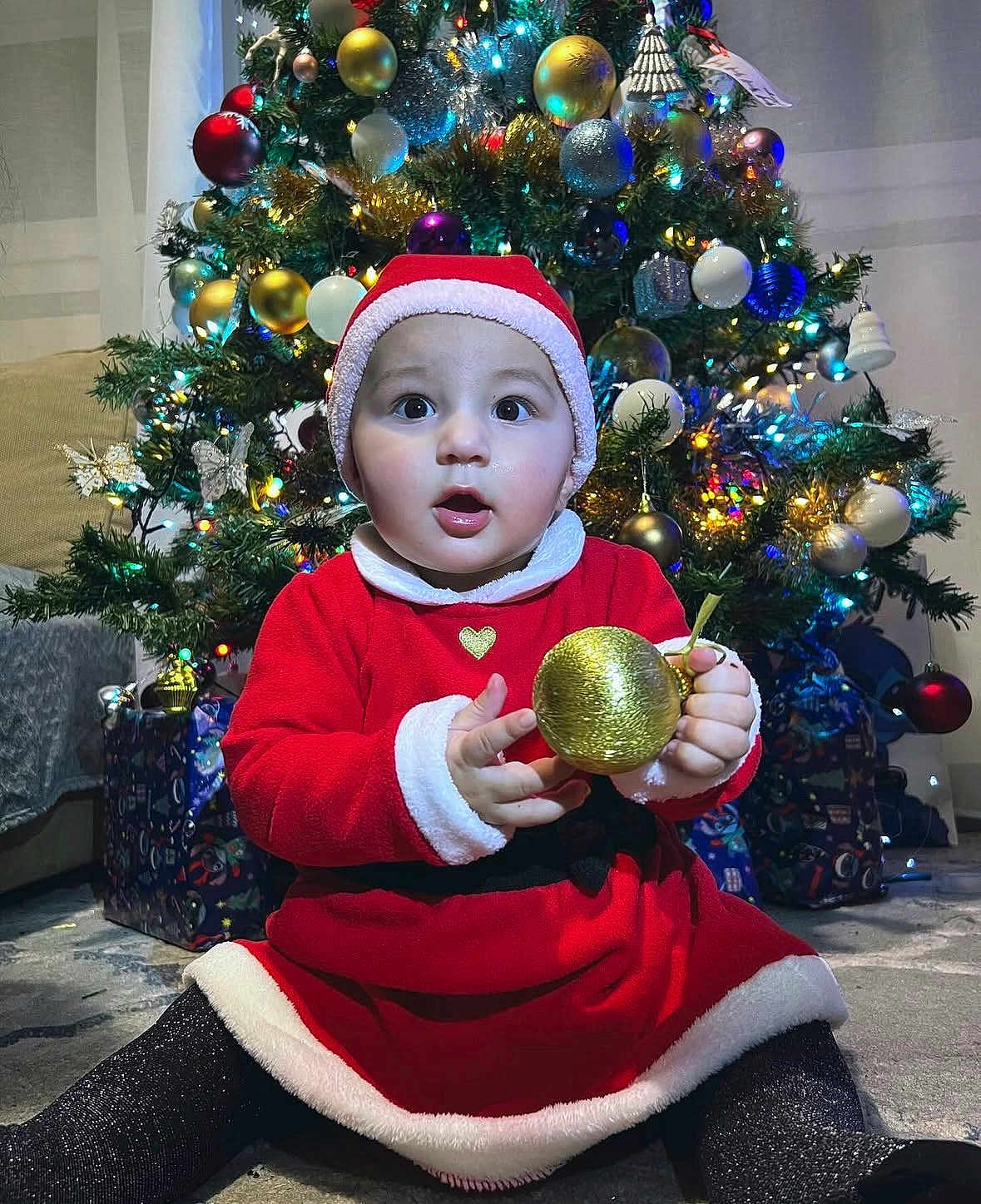 Esra a rejoint le concours — aidez-le/la à gagner de superbes lots ! baby, child, christmas_tree, ornament, holiday, festive, red_clothing, santa_hat, indoor, decorations, presents, sparkle, glitter, cute, celebration, seasonal, smiling, sitting, person, toy