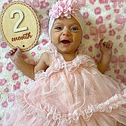 Novalina is registered to the contest to win money with this photo: baby, infant, smile, pink_dress, headband, flower, blanket, pattern, wooden_sign, milestone, happy, cute, portrait, celebration, lying_down, child, two_months, soft_texture, fabric, hands_up